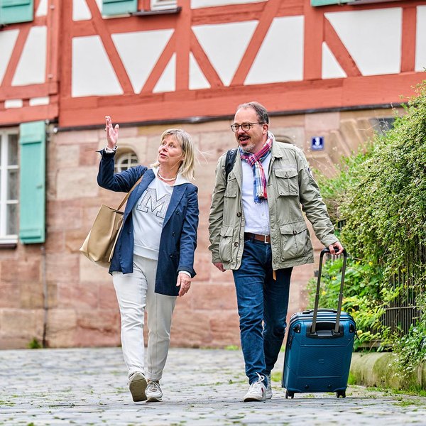 Zwei Best-Ager-Touristen mit Koffern laufen über den Kirchplatz in Fürth, hinter ihnen ein Fachwerkhaus.