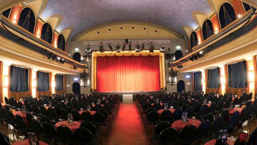 Heller Jugendstil-Theaterraum der Comödie Fürth mit Tischen, Stühlen und beleuchteter Bühne mit rotem Vorhang.