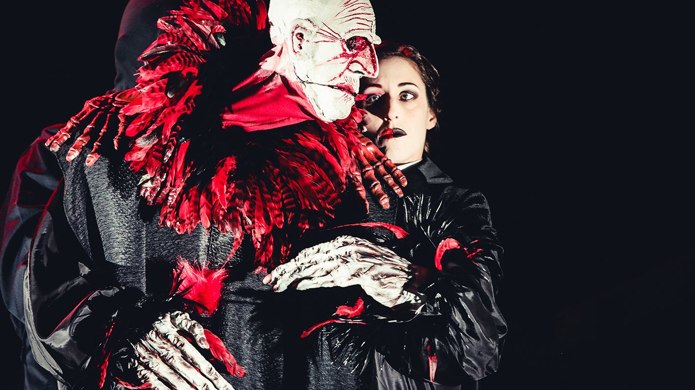 Große Puppe mit weißem Gesicht und roten Federn, geführt von einer Person in dunkler Kleidung.